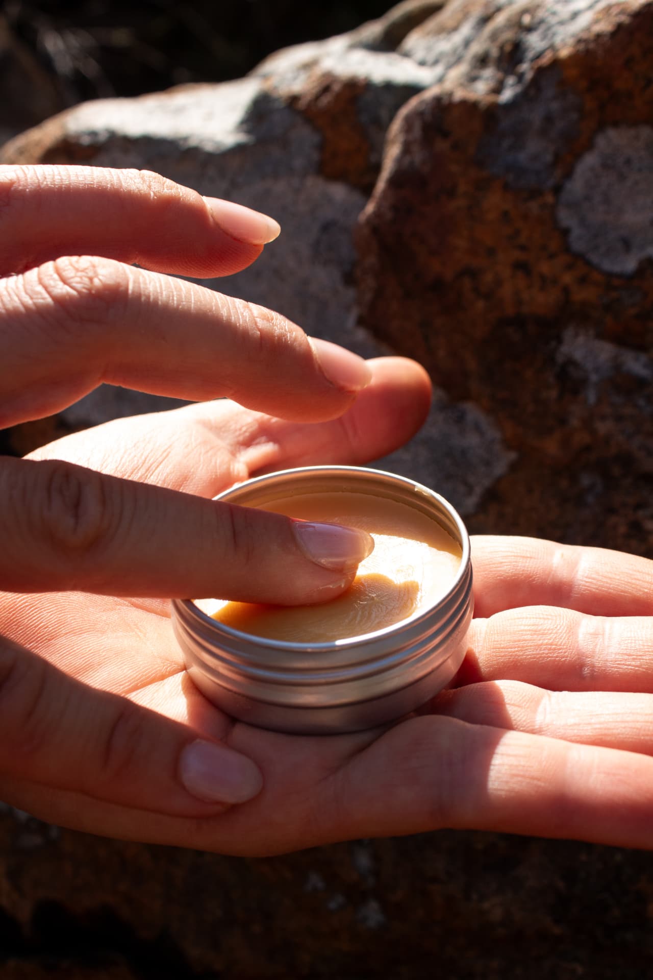 Fingers dipping into Everything Balm in an open tin held against sunlit lichen-covered rock
