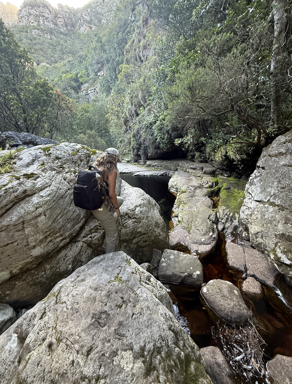 A walker stepping across sunlit boulders beside a dark mountain pool, a forested ravine rising behind