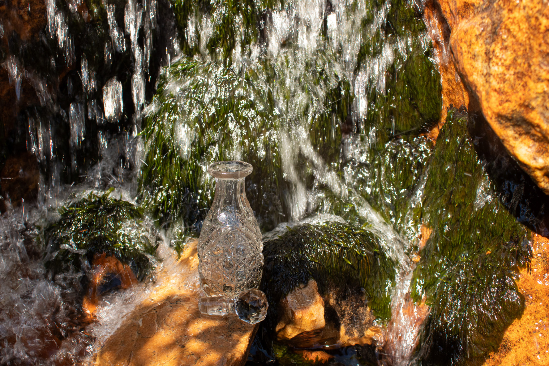 A cut-crystal decanter resting on sunlit rock beneath a mossy mountain cascade
