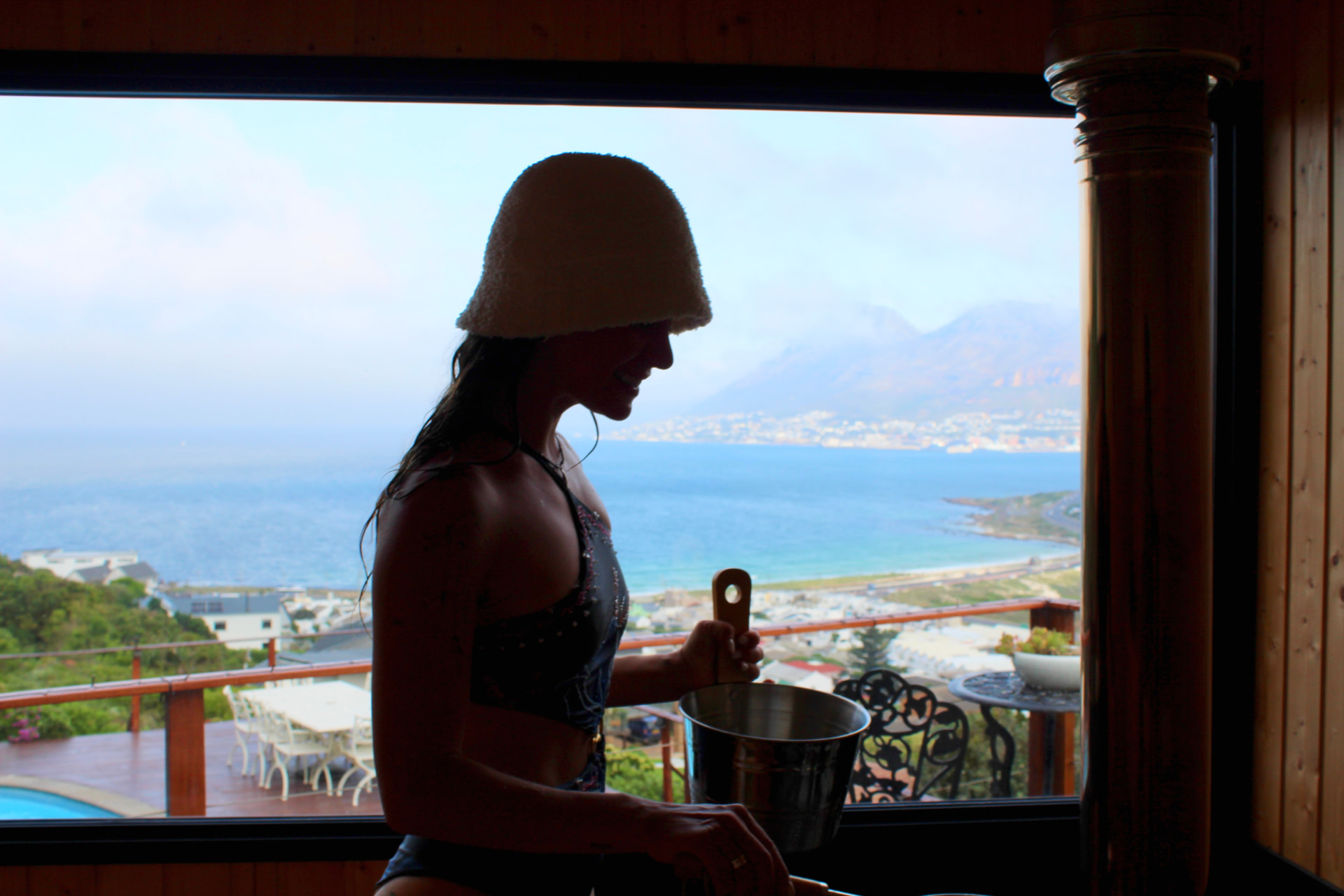 Mikaela in a white sauna hat holding a bucket inside a wood-fired sauna, looking out over a coastal bay