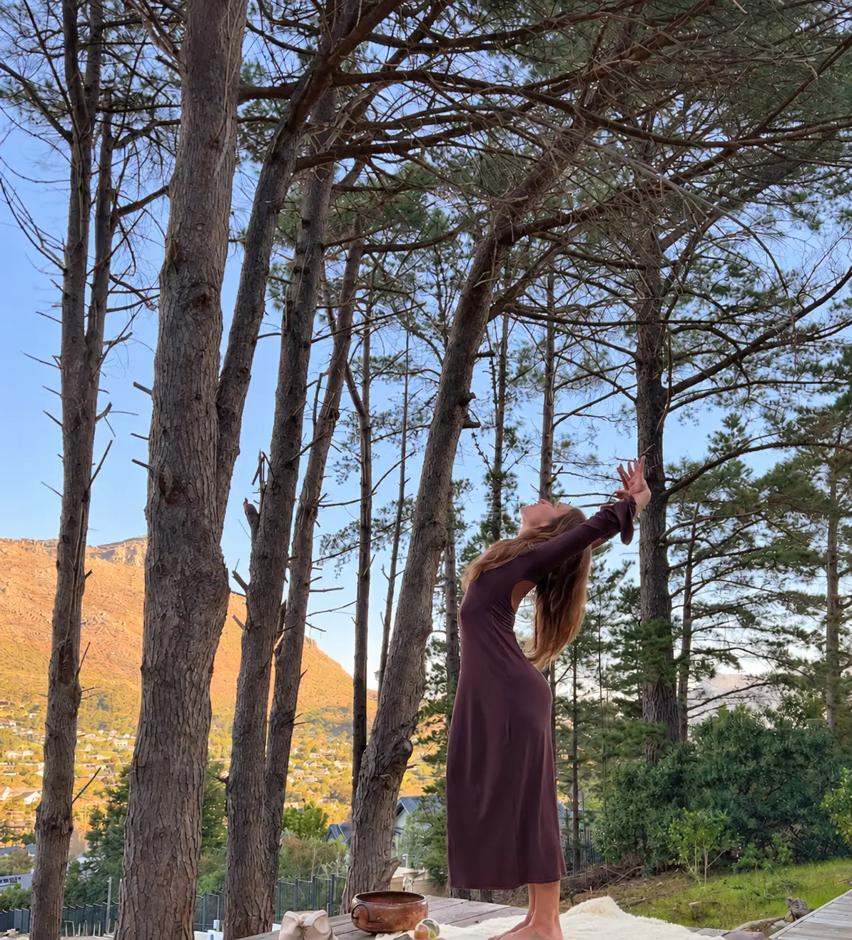 Mikaela arching back beneath tall trees on a wooden deck, mountains glowing in late afternoon light